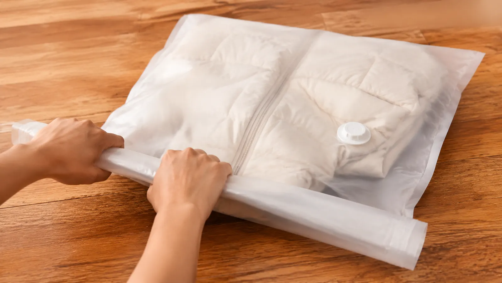 Hands rolling air out of a vacuum storage bag containing a folded down jacket on a wooden floor