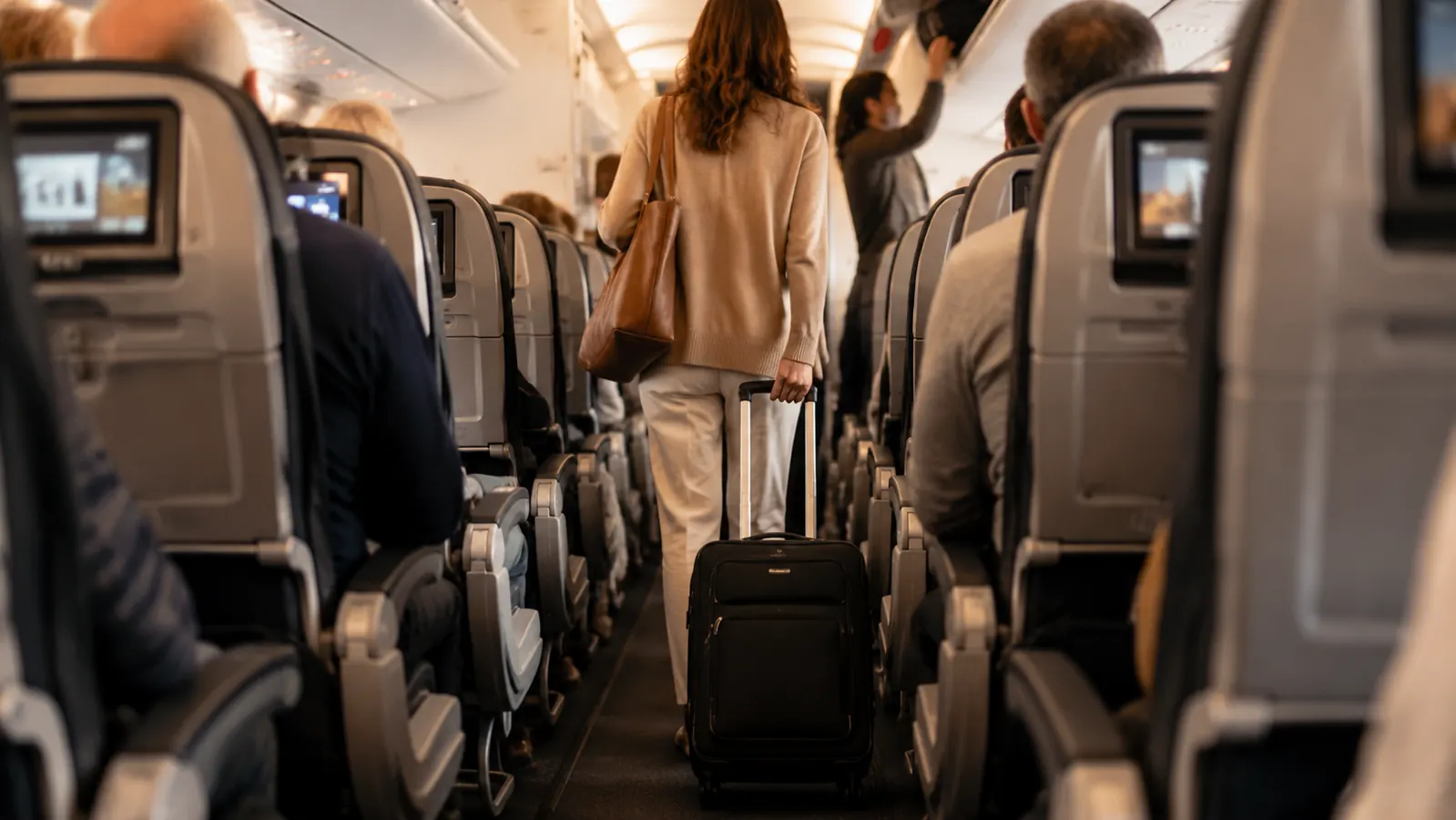 Woman pulling a black carry-on through a narrow airplane aisle with the bag fitting easily between seats